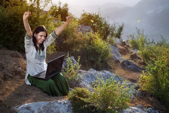 woman raising both arms while using her laptop in nature